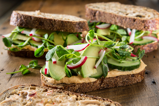 Three sandwich's on multigrain bread with red pepper hummus ,cucumber ,pea shoots ,avocado and radish's on a rustic wood background .