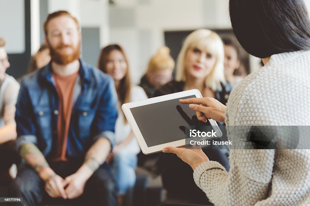 Teacher holding a digital tablet against auditorium Back view of woman holding a digital tablet and pointing with index finger at touchscreen. Group of smiling student in the background.  Digital Tablet Stock Photo