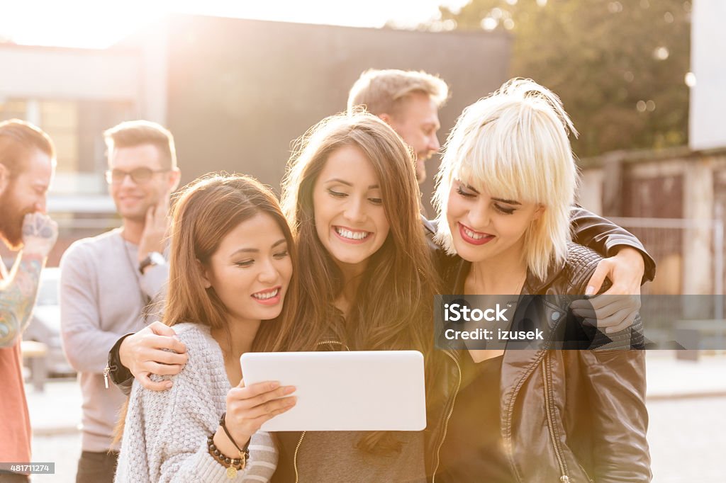Three grils taking selfie using a digital tablet Outdoor portrait of happy friends - three young women taking selfie using a digital tablet, guys in the background. 20-24 Years Stock Photo