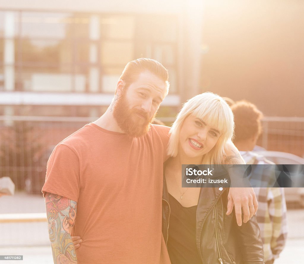 Contemporary couple - bearded guy with tattoos and blond girl Outdoor portrait of contemporary young couple in the city - redhair, bearded man with tattoos and blonde young woman wearing black leather jacket. Embracing and smiling at camera at sunset. 20-24 Years Stock Photo