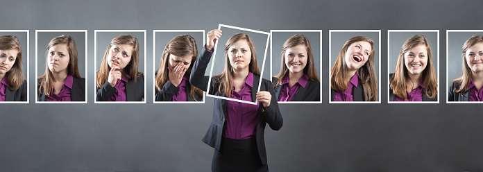 Concept photo for personality, character and emotional expression. A woman in business suit holding up a photo of herself in front of her with various range of emotional expressions exhibited in the background wall behind her. From happiness to sadness, anger to joy, apprehension to confidence. Photographed in panoramic horizontal format in studio.
