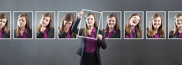 Concept photo for personality, character and emotional expression. A woman in business suit holding up a photo of herself in front of her with various range of emotional expressions exhibited in the background wall behind her. From happiness to sadness, anger to joy, apprehension to confidence. Photographed in panoramic horizontal format in studio.