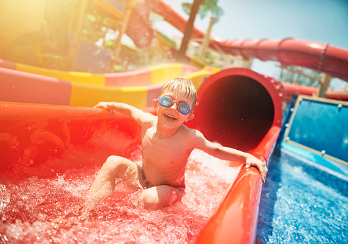 Little boy smiling into the camera after sliding on water slide in water park. The boy aged 5 is wearing swimming goggles and is laughing happily.