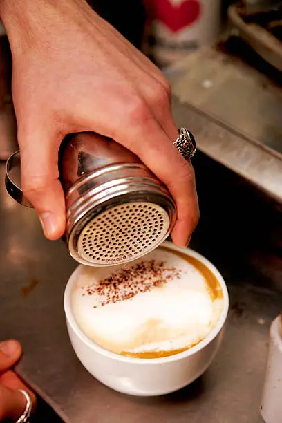 Close up of a barista's hand holding a silvery chocolate powder shaker over a fresh capuccino coffee.