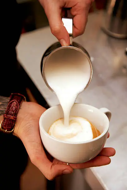 Close up of a barista's hand holding a white china cup with fresh coffee and pouring milk into it to make a cappucino.