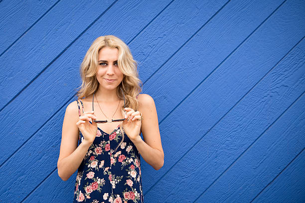 Pretty girl takes off sunglasses in front of blue wall stock photo