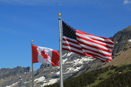 American and Canadian flags, showing the full stars and stripes and maple leaf respectively, on silver flagpoles blowing in the wind against a backdrop of snow covered mountains and forest trees