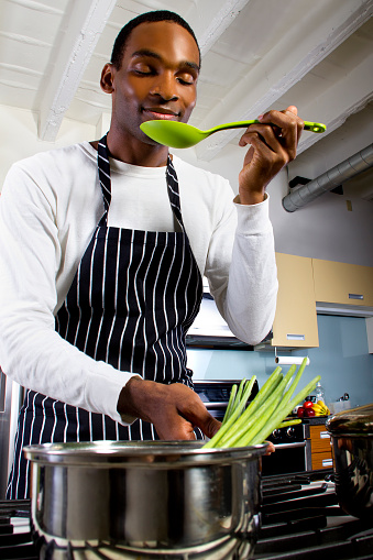 Close up photo of a young black man wearing an apron and cooking at home.  He is cooking vegetables in a pot on a stove.  He is in a domestic kitchen.  The man is preparing a healthy vegan meal.  The image depitcs diet and nutrition.
