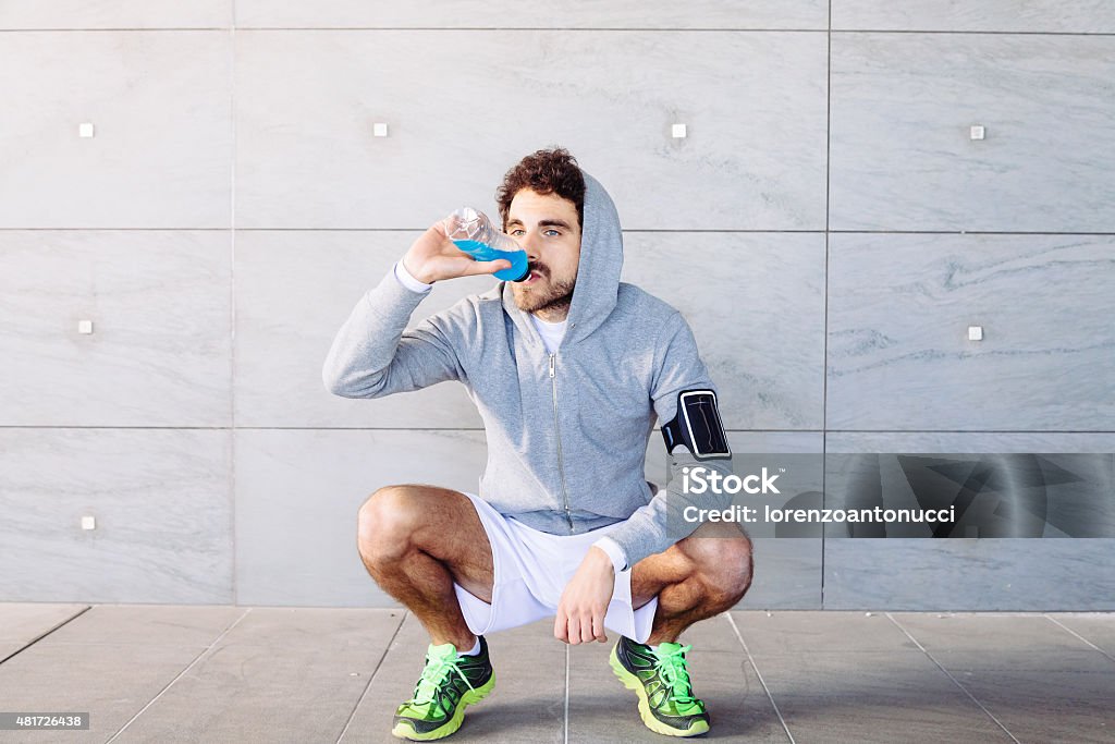 Young man resting on a run A young man in sportswear resting in the shade of a building in a summer day, drinking an energy drink blue color from a bottle before resuming training Drinking Stock Photo