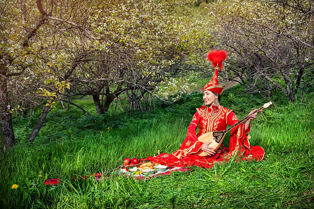 Kazakh woman in red costume playing dombra on the green grass in the apple garden of Almaty, Kazakhstan, Central Asia