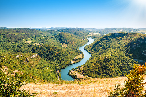 Horizontal composition photography of mountains, countryside rolling landscape of Ain river valley, with tree forest and arch bridge crossing the preserved natural Ain river. Image taken in Bugey, Rhone-Alpes region in France (Europe). This picture was made during a bright sunlight with the sun in the right top corner of the photo, and a clear blue sky without any clouds. Near to Alps chain and Jura in Franche Comte. Shot in middle of summer season with colorful tree and hill, with an impressive unobstructed view on rolling landscape mountain under sunlight.