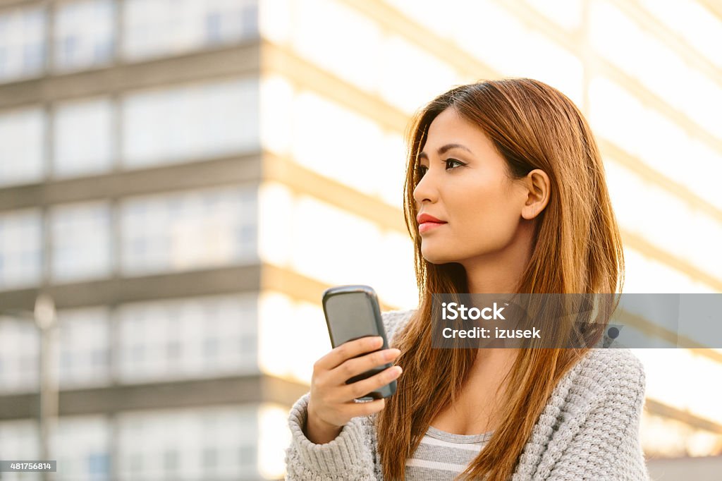 Asian young woman using smart phones outdoor Outdoor portrait of beautiful asian young woman using smart phones in the city at sunset, looking away, building in the background. 20-24 Years Stock Photo