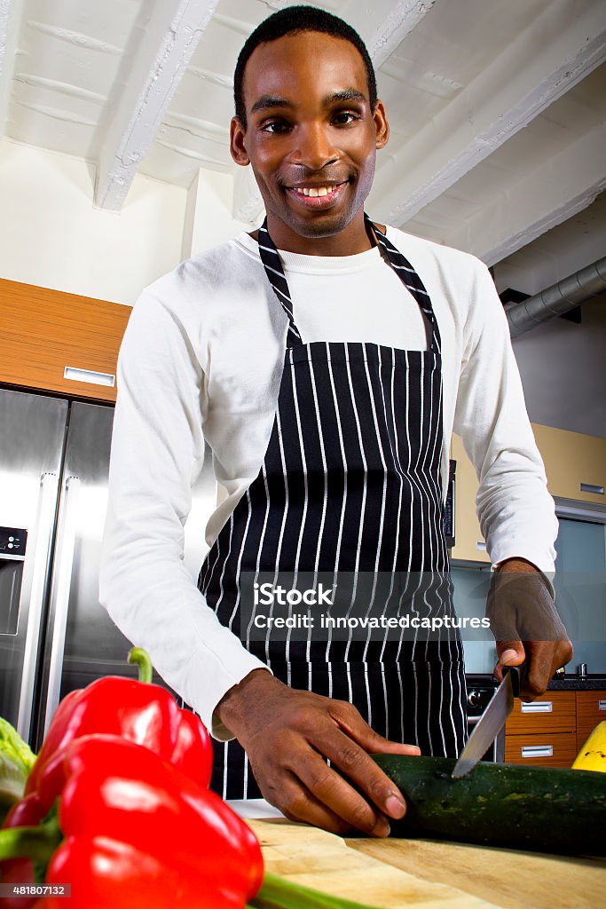 Cooking At Home Close up photo of a young black man wearing an apron and cooking at home.  He is chopping vegetables on a chopping board with a knife.  He is in a domestic kitchen.  The man is preparing a healthy vegan meal.  The image depitcs diet and nutrition. African Ethnicity Stock Photo