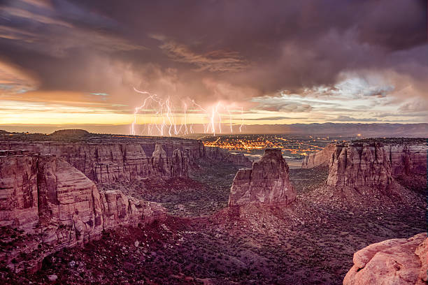 Independence Rock at Colorado National Monument during Sunset wi Dramatic lighting and clouds during sunset on the famous Colorado National Monument in Western Colorado near Grand Junction.  The feature is Independance Rock at the center of the image.  City lights of Fruita, Colorado are in the valley below. red rocks landscape stock pictures, royalty-free photos & images