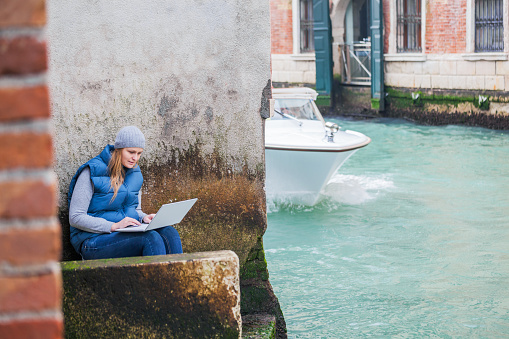 Young woman using laptop while sitting by the grungy wall near Venetian canal with sailing motor boat