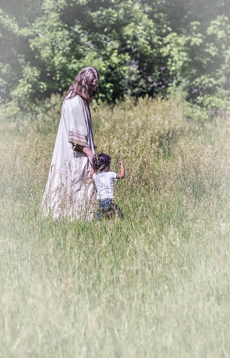 Jesus Christ is walking in a grassy meadow holding hands with a small three year old Hispanic boy. The child is gently touching the tall grass stalks as he walks and explores with Jesus. June 2015 Utah USA War and Peace photo shoot.