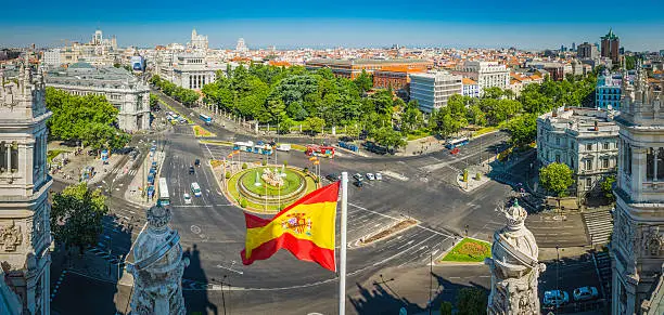 Spanish national flag flying proudly from the roof of the Palacio de Comunicaciones overlooking a panoramic vista across the busy Plaza de Cibeles towards the heart of central Madrid, Spain's vibrant capital city. ProPhoto RGB profile for maximum color fidelity and gamut.