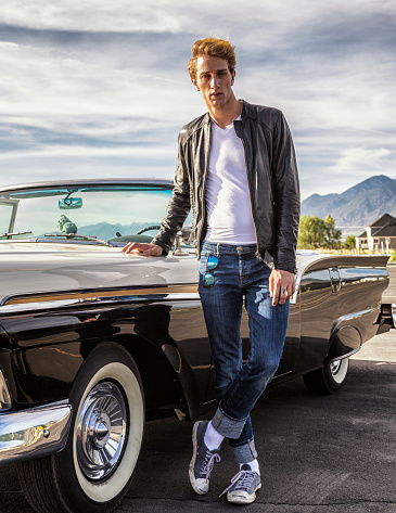 Back to the Fifties!! Tall, handsome Fifties greaser guy standing next to his hot convertible with the top down, and with iconic fuzzy dice hanging from the rear view mirror. He is wearing the 1950's young gun rebel uniform - white t-shirt, tight blue jeans, white socks with canvas sneakers, and a black leather jacket. It's a classic car and a classic pose by this classic young man who is clearly too cool for words. :) Utah War and Peace Photo Shoot - June 2015.