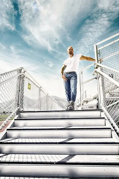 50's Tough guy standing alone in a stadium at the top of some steep metal stairs.  Big blue sky behind him with whispy clouds accenting the blue marine color in the sky behind him.