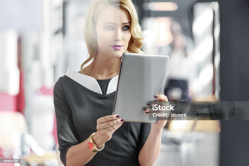 Portrait of an attractive businesswoman holding a digital tablet Portrait of beautiful blonde young businesswoman wearing grey dress sitting in an office and holding a digital tablet, looking away. Photo taken through glass. 2015 Stock Photo