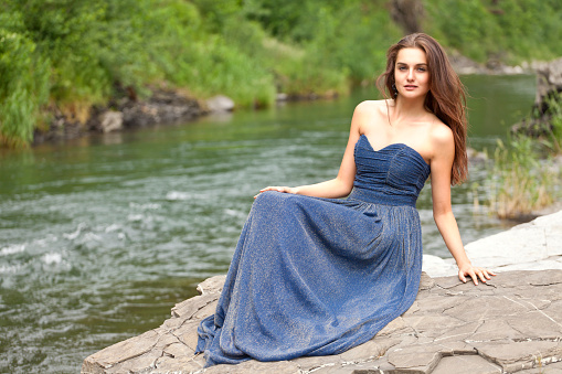 Young attractive brunette in long blue dress is sitting on a rock by the river. Blurred background of the river and green nature, she is smiling and looking at the camera. Summer atmosphere. Shooting with Canon EOS 5D Mark II.
