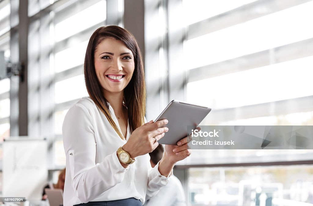 Businesswoman using a digital tablet in an office Friendly businesswoman wearing white shirt sitting in a board room in an office, holding a digital tablet in hands and smiling at camera.  Businesswoman Stock Photo