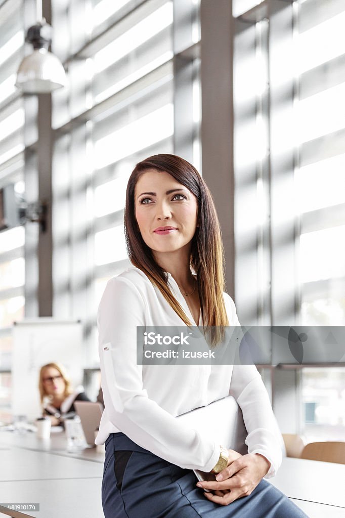 Attractive businesswoman in an office, holding a digital tablet Friendly businesswoman wearing white shirt sitting in a board room in an office, holding a digital tablet in hands, looking away. 2015 Stock Photo