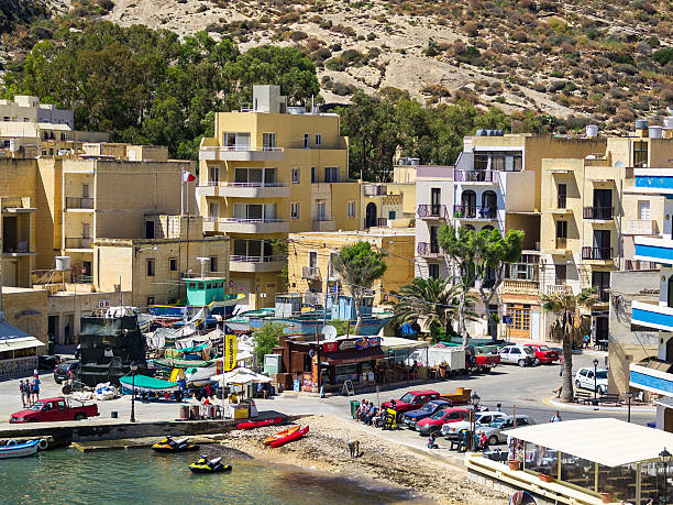 Marsalforn, Malta - May 29, 2015: Horizontal view of small town of Marsalforn on Island Gozo. It is a small harbor with boats in water or parked on land. There are many cars parked on harbor square and some tourists relaxing on bench near water not far from restaurant and  store on other side of square. In background are town buildings and rocky hill.