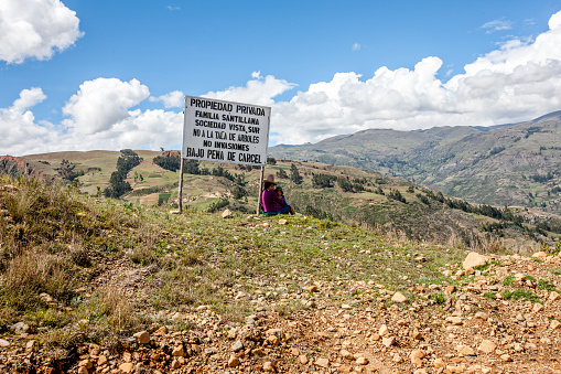 Huaraz, Peru - January 24, 2015: Campesina mother and her child in high Andes sitting under a sign while looking after her donkeys grazing on the hillside