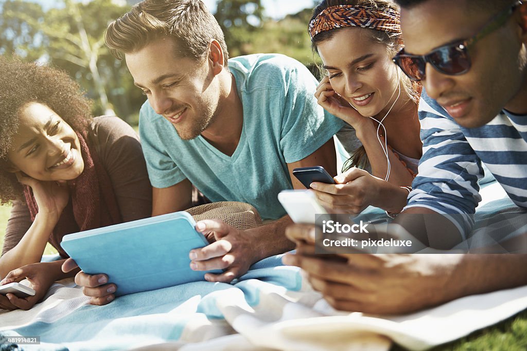 Never far from their toys Shot of a group of young people using their phones and a digital tablet at the park 20-29 Years Stock Photo