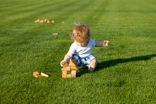 Little curly baby boy sat down to build toy wooden house on the green lawn sunny day outdoor, horizontal photo