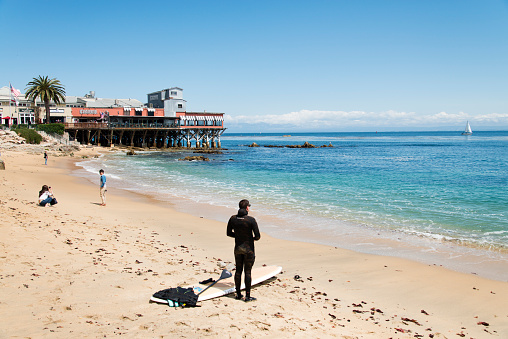 Monterey, California, USA - May 15, 2015: View at a beach in the city of Monterey during a clear summer day. A surfer standing on the beach while some other people are walking and sitting in the background. In the background also some restaurants are showing.