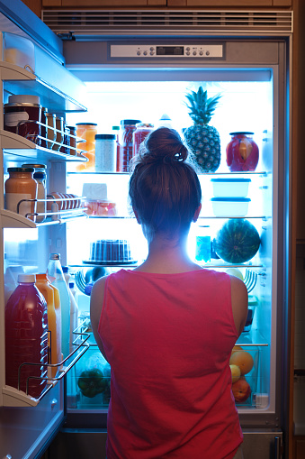 A young caucasian woman standing in front of the open refrigerator at late night, contemplating and wondering about a midnight snack in a domestic home kitchen. She is dressed in a bath robe hungry and looking for food. A symbol of dieting lifestyle. Photographed in vertical format.