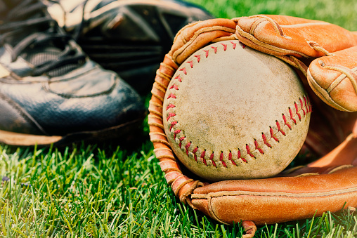 A Vintage looking image of a close-up of an old softball in a leather glove with black cleats in the grass as the sun shines down. A perfect day for a softball game in the park.