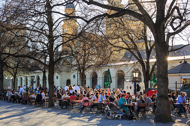Munich, Germany - March 20, 2014: People enjoying the early spring sun in an outdoor cafe and beer garden in the park Hofgarten in the center of Munich. In the background the Theatine church. The cafe is called Tambosi.