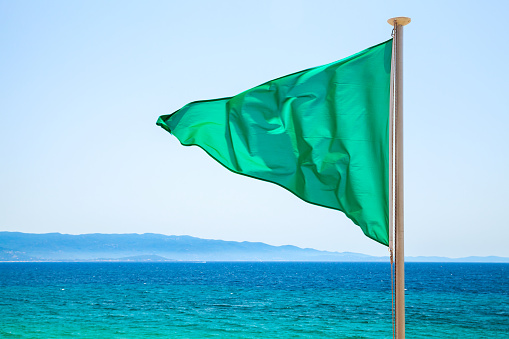 Green flag on the beach over bright blue sea background, means that swimming is allowed