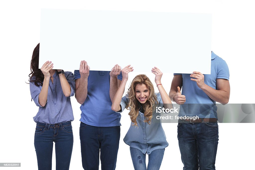 smiling woman holding banner with her friends hiding casual people hiding behind a blank board, one smiling woman looks to the camera and one man making the ok thumbs up sign Admiration Stock Photo