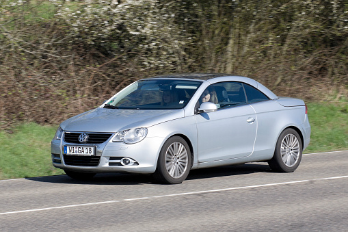 Near Wiesbaden, Germany - April 2, 2011: Two women in an accelerating silver-metallic Volkswagen EOS (2-door coupe convertible) on a country-road in the Rheingau (Germany) on a sunny spring day. The VW Eos (Typ 1F) is a four-seat coupe convertible with retractable metal hardtop. It was introduced in 2006. VW is a brand of the Volkswagen Group, which is a German automobile manufacturing group based in Wolfsburg, Germany and founded in 1937. Some motion blur.