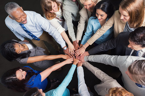 Team of diverse business professionals are standing in a circle in their office. They are placing their hands in a huddle circle to celebrate success or strategize teamwork.  Senior adults, mature adults, mid adults, and young adults are part of large professional group. View from above shows team of people wearing professional business casual clothing.