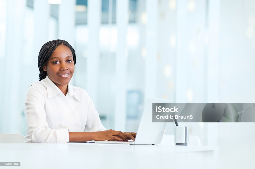 Smiling African American businesswoman using laptop in the office. Young smiling African American businesswoman sitting in the office and using computer. She is looking at the camera. 2015 Stock Photo