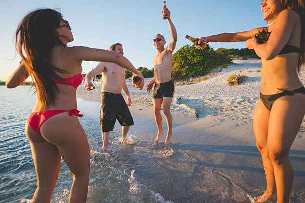 group of young multiethnic friends women and men at the beach in summertime toasting and drinking beer jumping and dancing - friendship, carefreeness concept