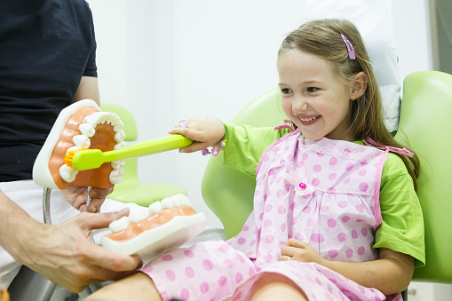 Smiling little girl in dentists chair, being educated about proper tooth-brushing by her paediatric dentist. Early prevention, raising awareness, oral hygiene demonstration concept.