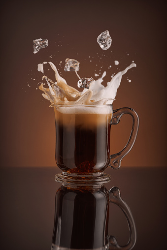 Splash of  ice coffee drink on a brown background. Refreshing Iced cappuccino liquid drink pouring into a mug, cup with ice cubes. Cold beverage wave. Close-up design liquor milk, coffee and ice. 