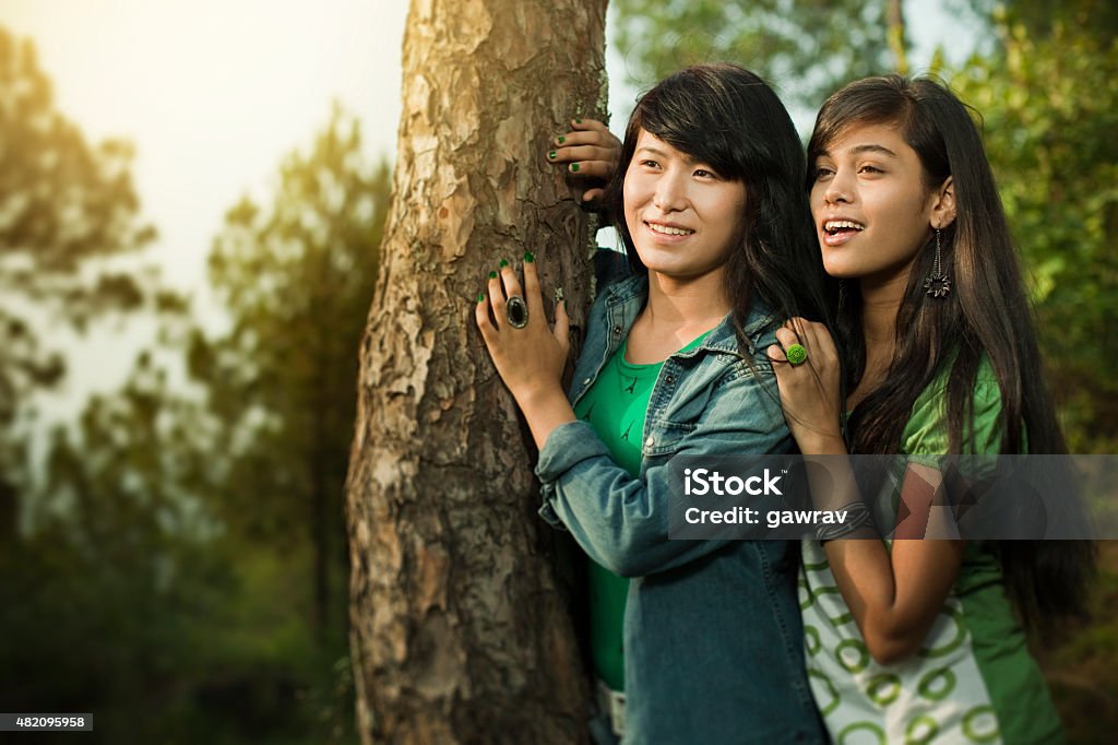 Two happy teenage girls from different ethnicity together in nature. Outdoor day time image of two Asian happy teenage friends (girls only) from different ethnicity. They are standing together by a tree trunk in rural natural environment and looking away at something wearing casual dress and giving toothy smile. Horizontal image with selective focus and copy space at aperture f/2.8. 16-17 Years Stock Photo