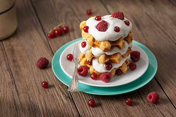 High resolution digital capture of a stack of golden, fresh, crispy, tender, Belgian waffles; layered with fresh whipped cream and adorned with fresh raspberries and lingonberries.  Waffles are stacked on a white plate sitting on a blue plate, on a rustic wooden table, with a sterling silver fork.