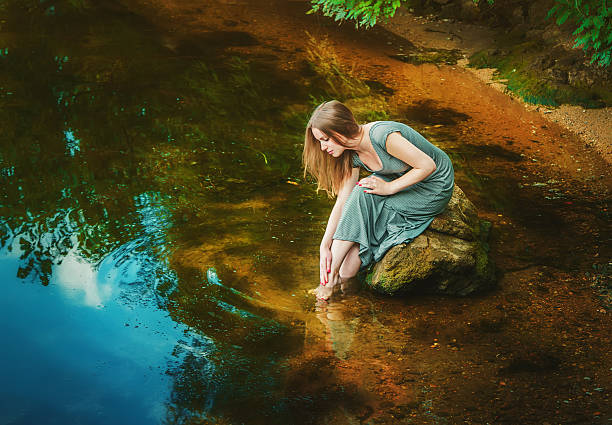 Attractive young woman wearing long dress sitting on the rock touching her feet in a weedy pond water
