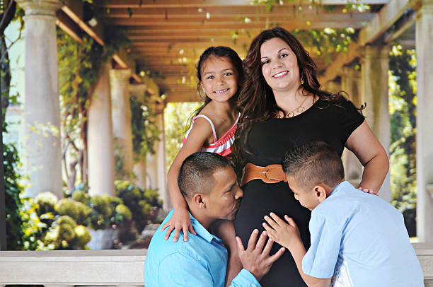 A happy  mom-to-be-again and her young daughter with the dad and elementary son touching and kissing her pregnant belly.