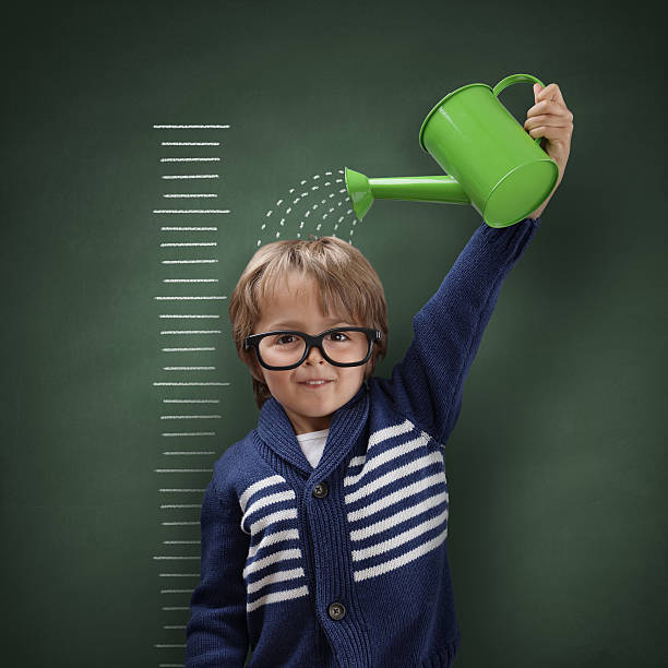 Growing up Young boy trying to make himself taller with watering can measuring his growth in height against a blackboard scale meter instrument of measurement photos stock pictures, royalty-free photos & images
