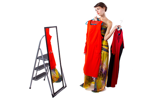Photo of a young female choosing clothes in a fitting room with a mirror.  She is isolated on a white background.  The woman is shopping for a colorful dress.  She is choosing a dress that fits and looks stylish and fashionable.  She is holding multiple dresses and being undecisive. The image depicts the fashion and clothing industry.