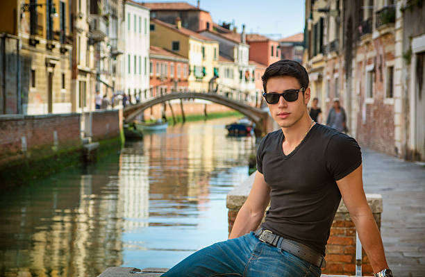 Portrait of Attractive Dark Haired Young Man Leaning Against Railing on Foot Bridge Over Narrow Canal in Venice, Italy